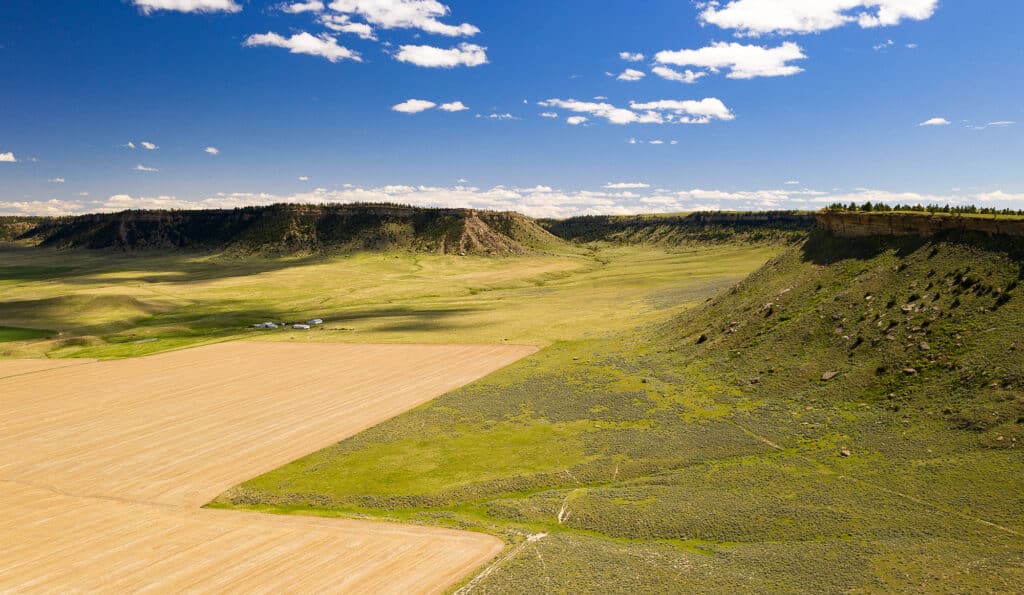 A scenic aerial view of recreational land for sale, featuring flat farmland, green fields, and steep rocky cliffs under a bright blue sky with scattered white clouds.