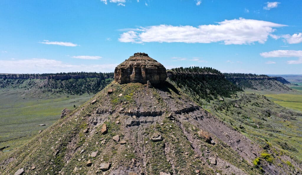 A rocky, dome-shaped butte rises from a sloping hillside in a vast, green landscape under a bright blue sky with scattered clouds. Sparse trees line the horizon atop the plateau, making this an ideal recreational land or hunting property.