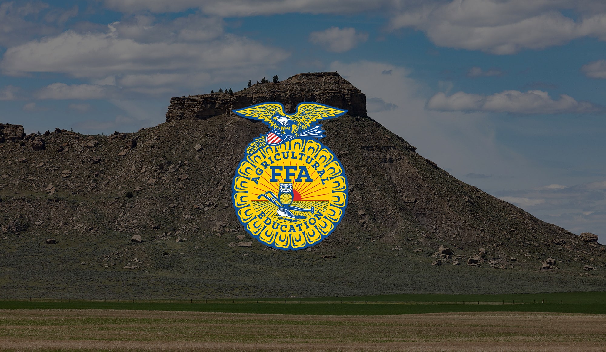 A rocky mesa rises above a field under a cloudy sky, with a large, bright National FFA Organization emblem digitally placed over the hillside—an ideal setting for recreational land or hunting property.