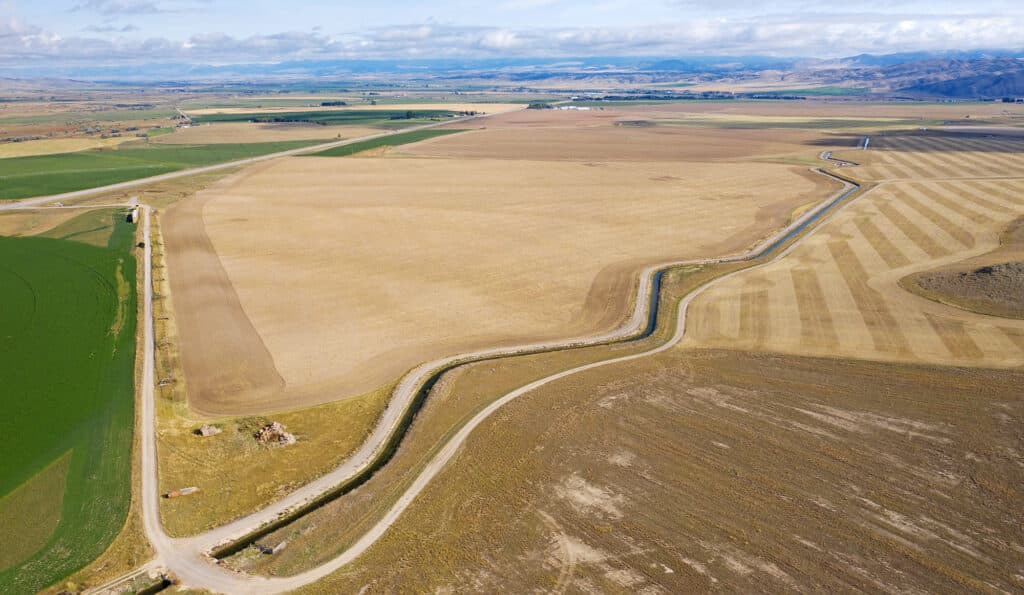 Aerial view of a rural landscape with large, patchwork fields—ideal as a cattle ranch or hunting property—divided by a winding dirt road under a partly cloudy sky. Mountains are visible in the distant background, perfect land for sale.