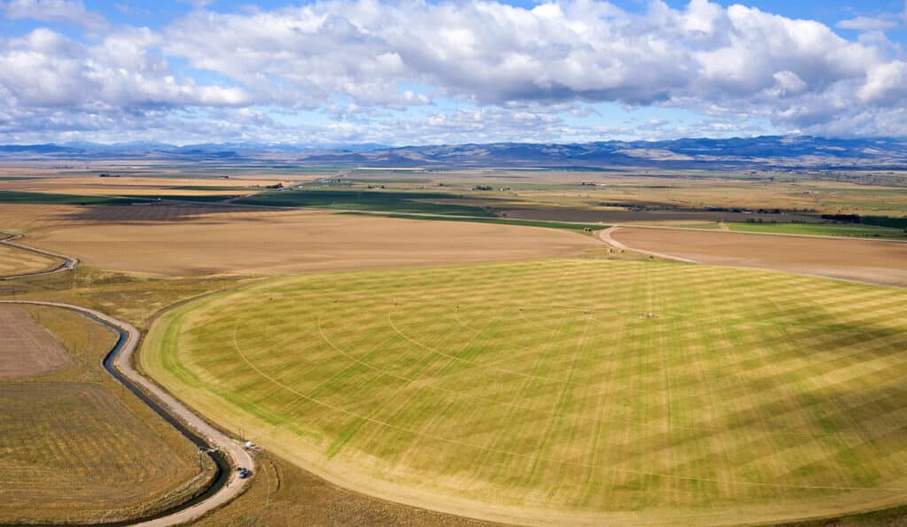 Aerial view of vast agricultural fields with geometric patterns, intersecting dirt roads, and irrigation lines under a partly cloudy sky; ideal as cattle ranch or recreational land, with distant mountains visible on the horizon.