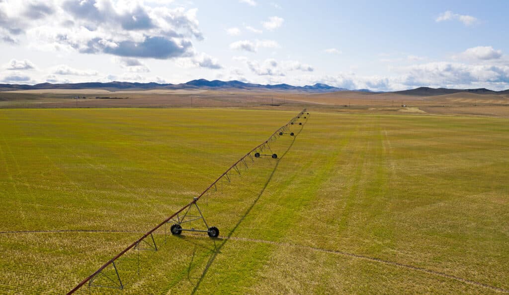 Aerial view of a large, green agricultural field with a long center pivot irrigation system; ideal cattle ranch land for sale, with mountains and a partly cloudy sky in the background.