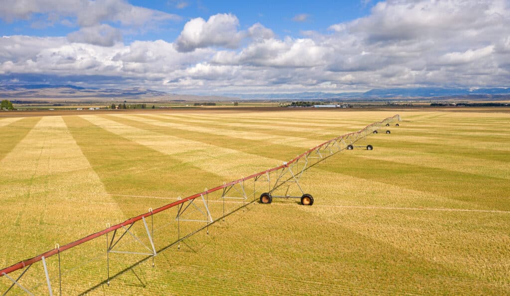 Aerial view of a large, flat agricultural field on recreational land with a long center-pivot irrigation system and scattered clouds in a blue sky above distant mountains.