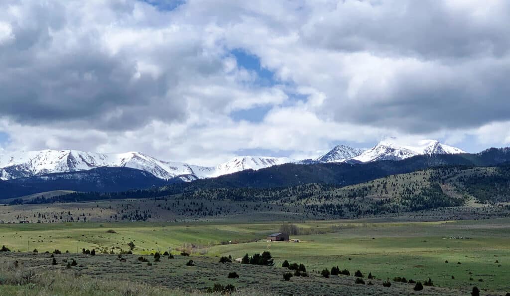 Snow-capped mountains under a cloudy sky rise above rolling green hills and fields, with a small building visible in the wide open landscape—perfect recreational land or hunting property.