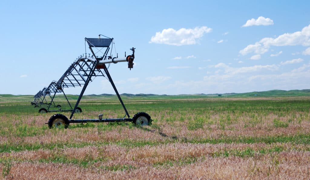 A large metal irrigation system stands on wheels in a flat, grassy field under a blue sky with scattered clouds. This open landscape stretches into the distance, making it ideal as recreational land or for a cattle ranch.
