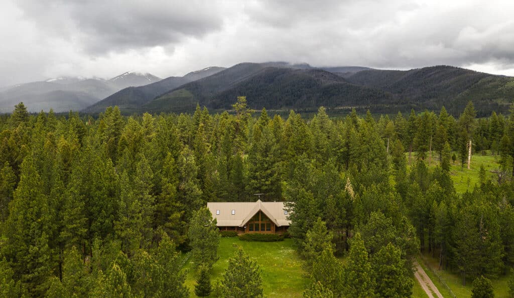 A wooden cabin on recreational land is surrounded by dense green pine trees in a clearing, with tall forested mountains and cloudy skies in the background.