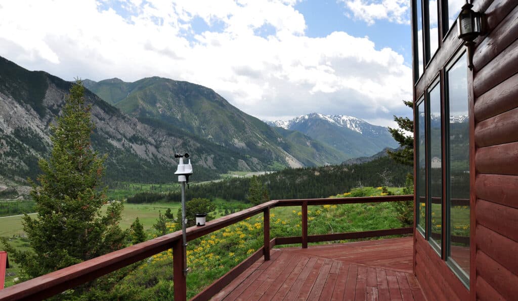 A wooden deck attached to a rustic cabin overlooks a lush green valley on hunting property, with tall pine trees and dramatic mountain peaks in the background under a partly cloudy sky.