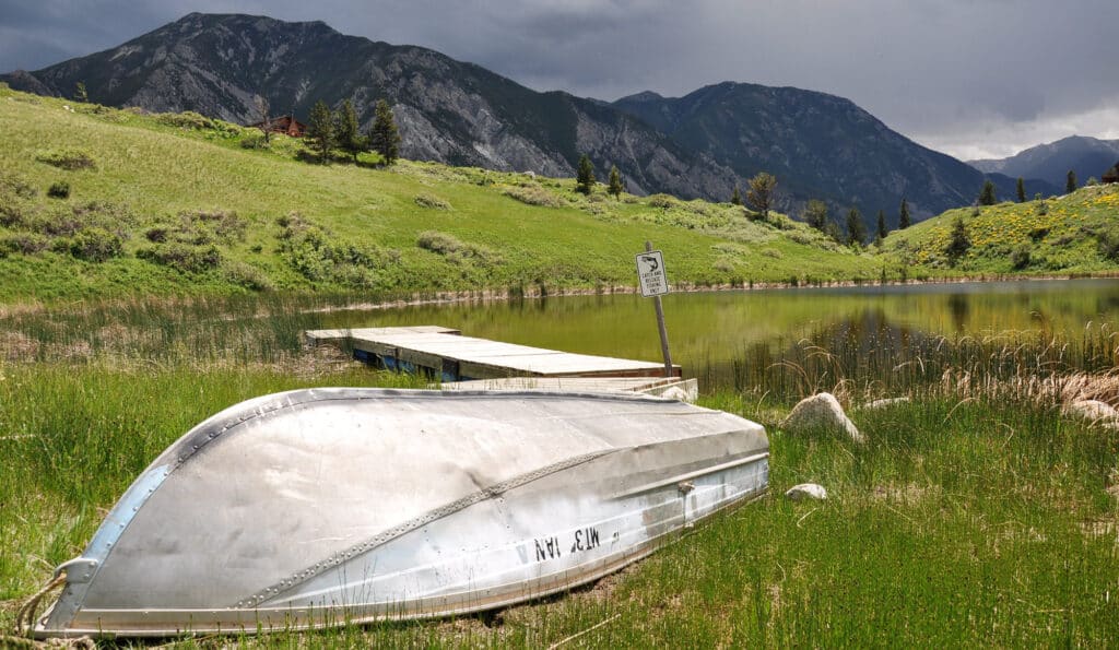 An overturned metal boat rests on grassy recreational land beside a small dock extending into a calm lake, with green hills and tall mountains in the background under a cloudy sky. A sign stands near the dock’s edge.