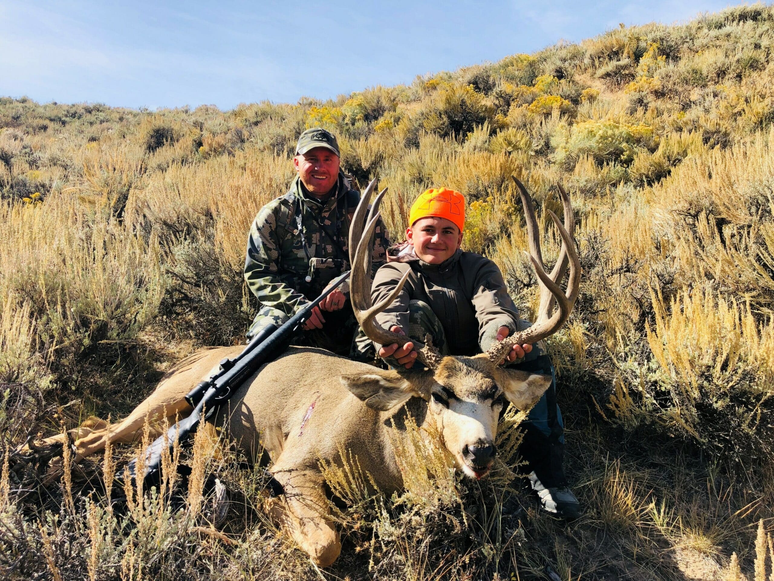 Two hunters pose outdoors on a hillside with dried grass and bushes. One holds a rifle and the other, wearing an orange hat, holds up the antlers of a large deer—highlighting the appeal of quality hunting property and recreational land.