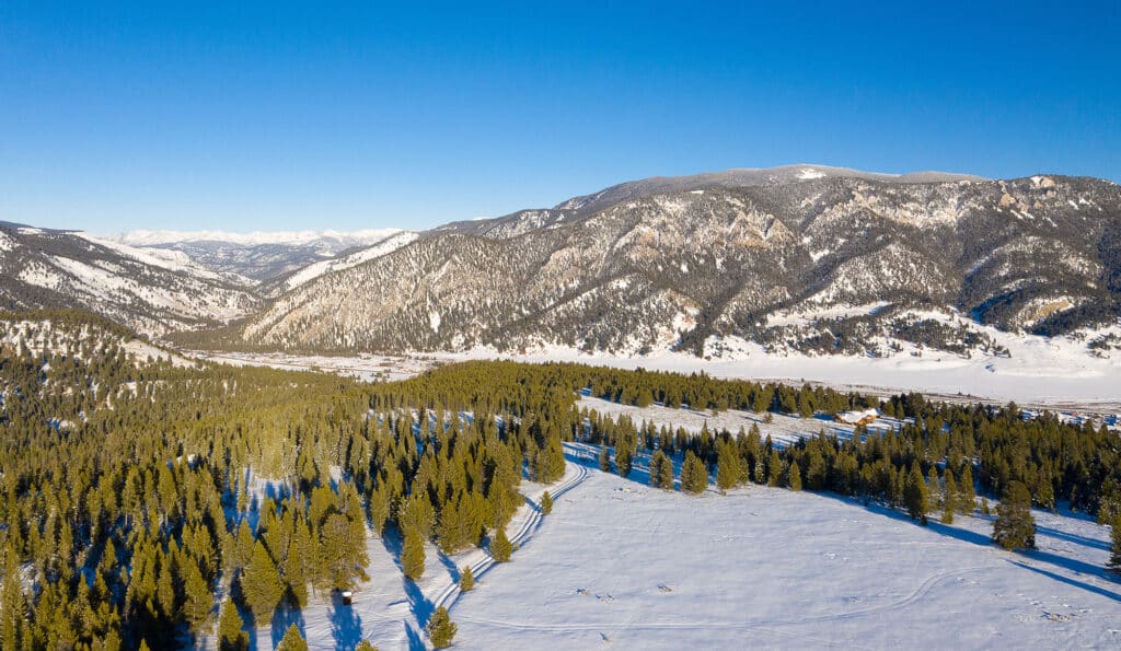 Aerial view of a snow-covered valley with scattered evergreen trees, surrounded by forested mountains under a clear blue sky—ideal recreational land or hunting property.