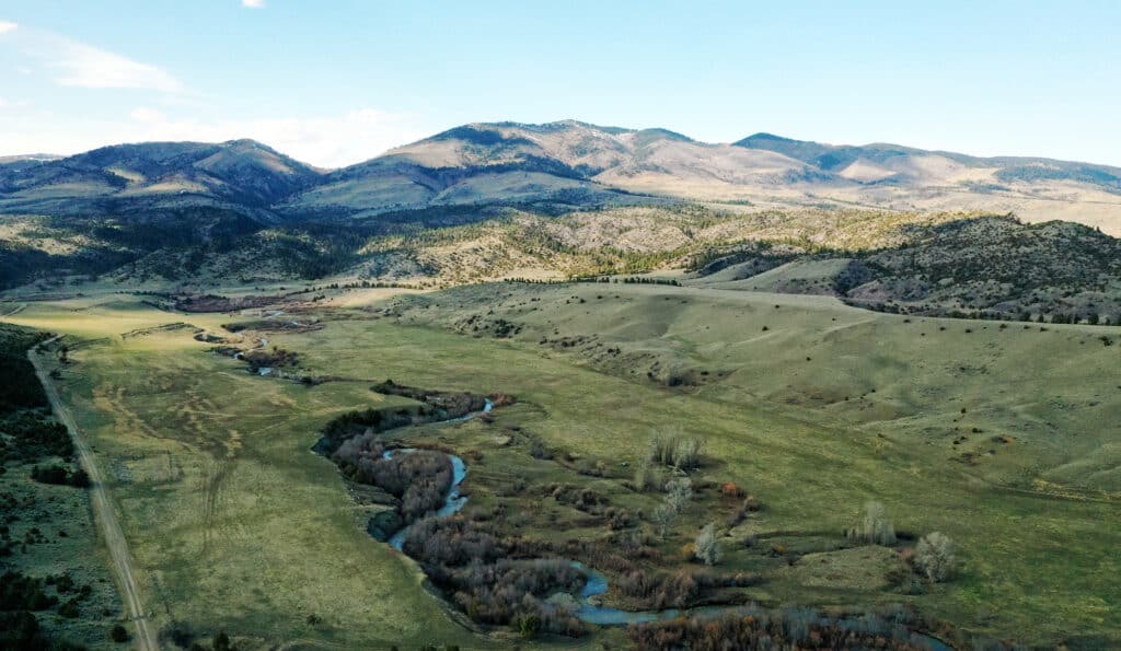 A scenic landscape featuring recreational land with a winding river flowing through a wide green valley, surrounded by rolling hills and mountain ranges under a bright blue sky.