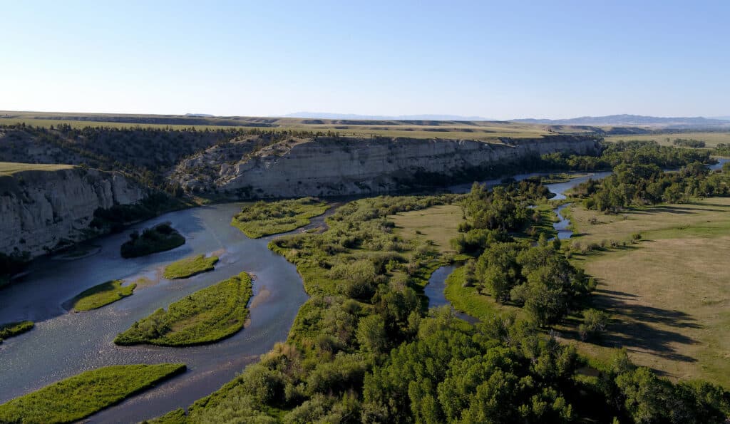 A wide river winds through a green, tree-lined valley with rocky cliffs and flat grasslands under a clear blue sky—ideal recreational land or potential cattle ranch.