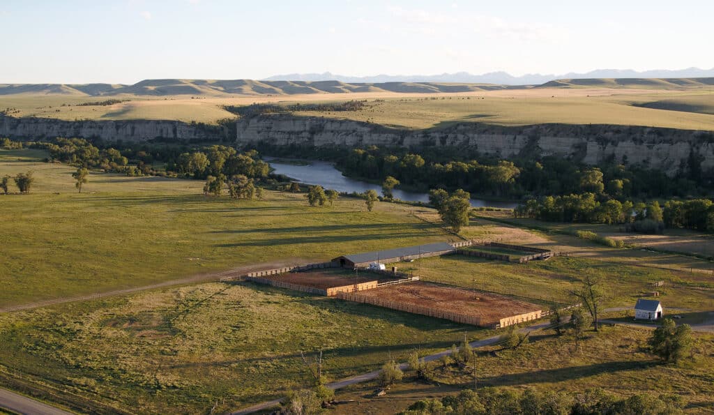 Aerial view of a rural landscape featuring a fenced cattle ranch with a barn and small white building near a river, surrounded by green fields, trees, cliffs, and distant hills under a clear sky.