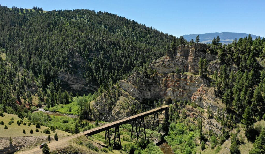 A wooden trestle bridge crosses a deep, forested canyon on a scenic cattle ranch, with rocky cliffs, green trees, and rolling hills framed by distant mountains under a clear blue sky.
