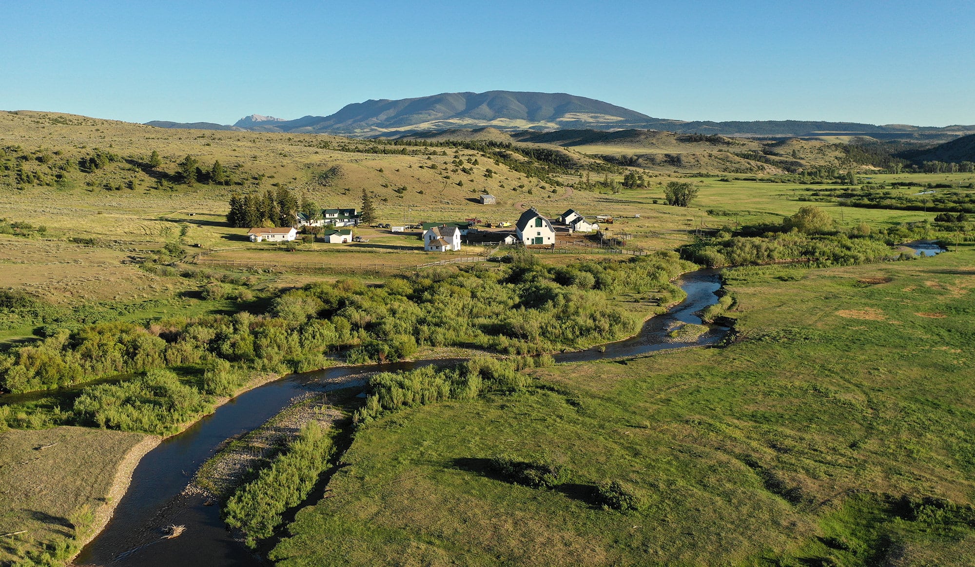 A winding river flows through a green valley with scattered houses and trees, surrounded by rolling hills and distant mountains under a clear blue sky—perfect recreational land or hunting property for nature lovers.