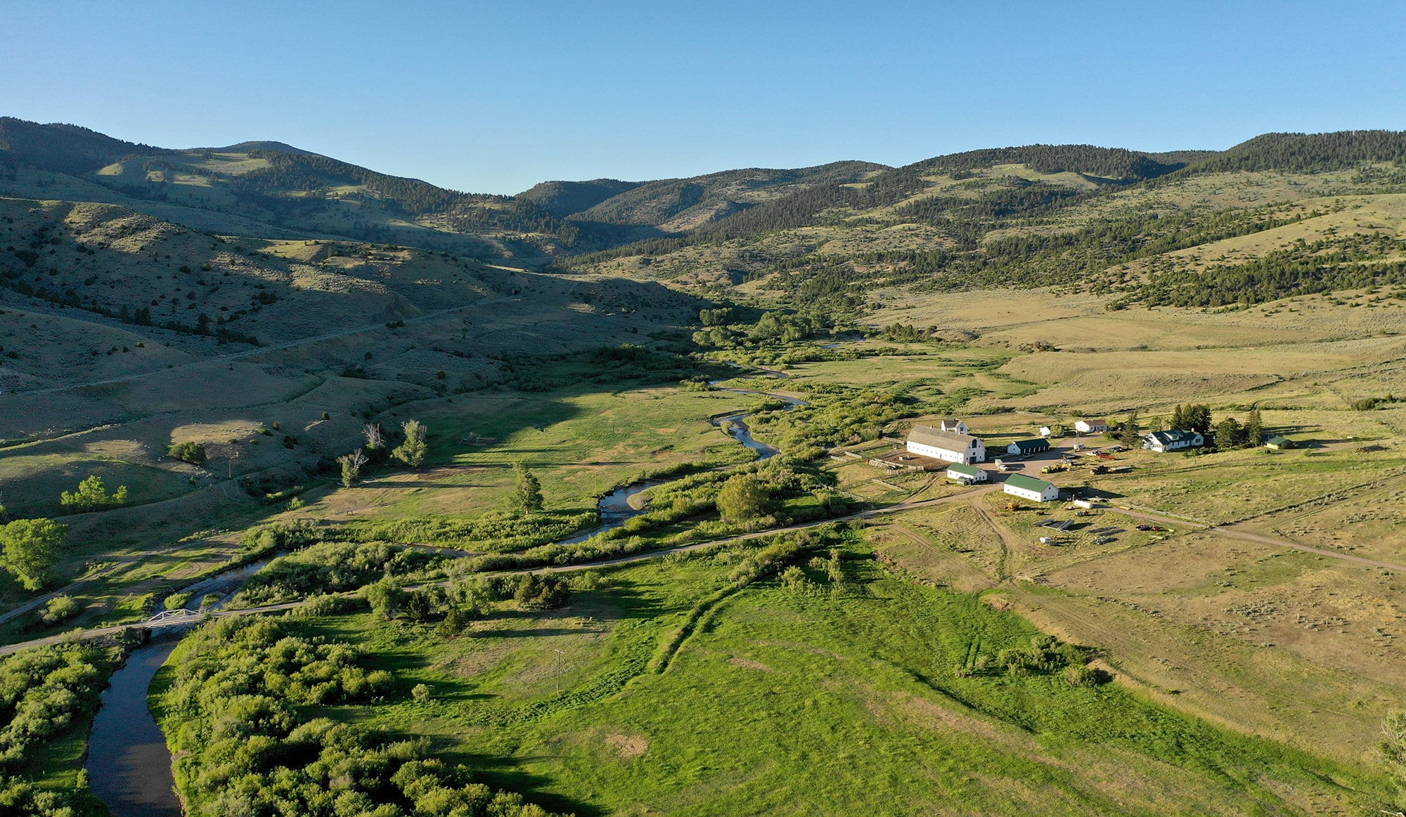 Aerial view of a rural valley with a winding river, green fields, scattered trees, and a cluster of farm buildings near gently rolling hills under a clear blue sky—ideal recreational land or cattle ranch for sale.