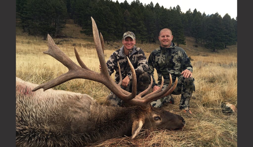 Two people in camouflage pose outdoors in a grassy field beside a large elk with antlers lying on the ground, showcasing the possibilities of this hunting property surrounded by pine trees and rolling hills.