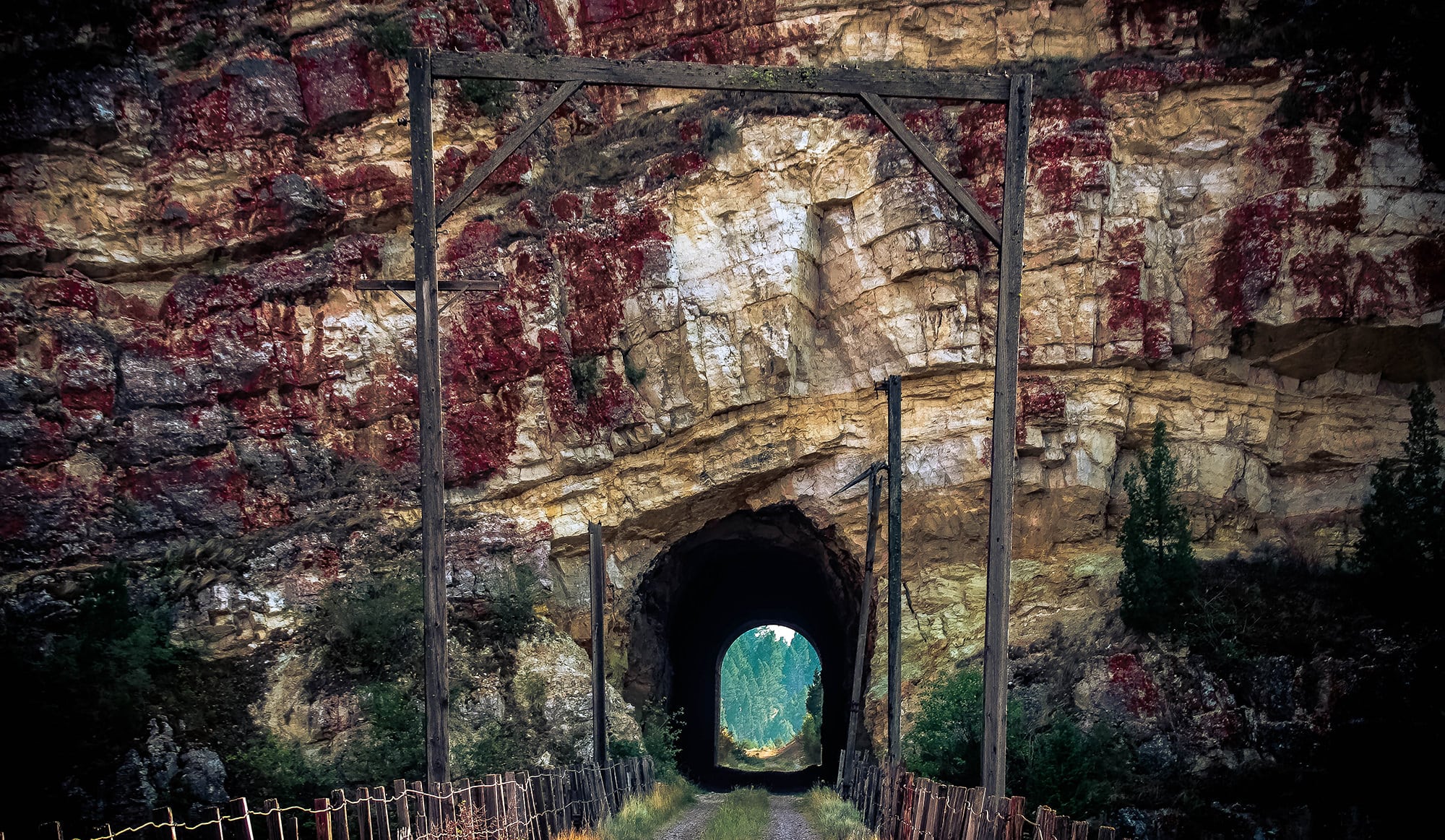 A dirt path lined with wire fencing leads to a rocky tunnel carved into a cliff—ideal for recreational land seekers. Light shines through the tunnel, revealing trees and greenery at its far end.