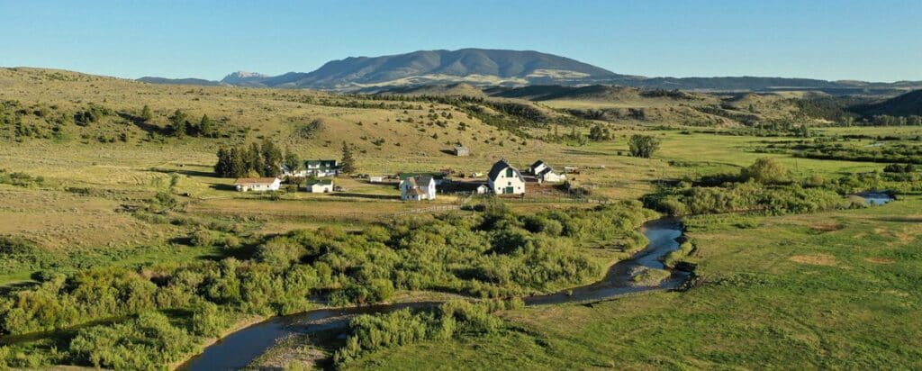 A small rural settlement with white buildings sits in a green, open landscape, ideal as a cattle ranch or recreational land, surrounded by grassy hills and a winding stream, with mountains visible in the distance under a clear blue sky.
