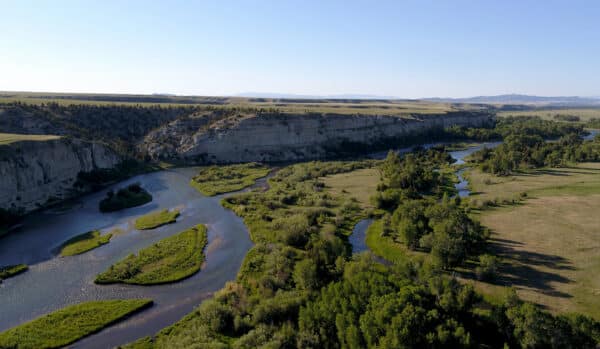 A winding river flows through a green valley with scattered trees, bordered by high, rocky cliffs under a clear blue sky. This expansive landscape in the sunlight presents an ideal recreational land or hunting property.