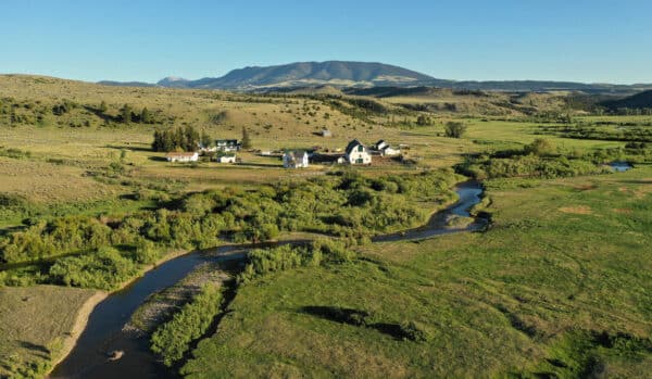Aerial view of a rural landscape with a small cluster of white houses and barns near a winding stream, surrounded by green fields and hills—ideal recreational land with distant mountains under a clear blue sky.