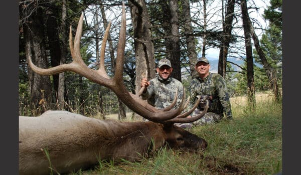 Two people in camouflage sit in a forest on hunting property next to a large elk with impressive antlers lying on the grass. Trees and distant hills create a scenic backdrop.