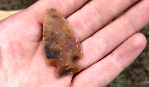 A hand holds a brown, translucent arrowhead made of stone, showing detailed flaking marks and a notched base. Blurred grass hints at open fields, evoking the spirit of land for sale or a cattle ranch in the distance.