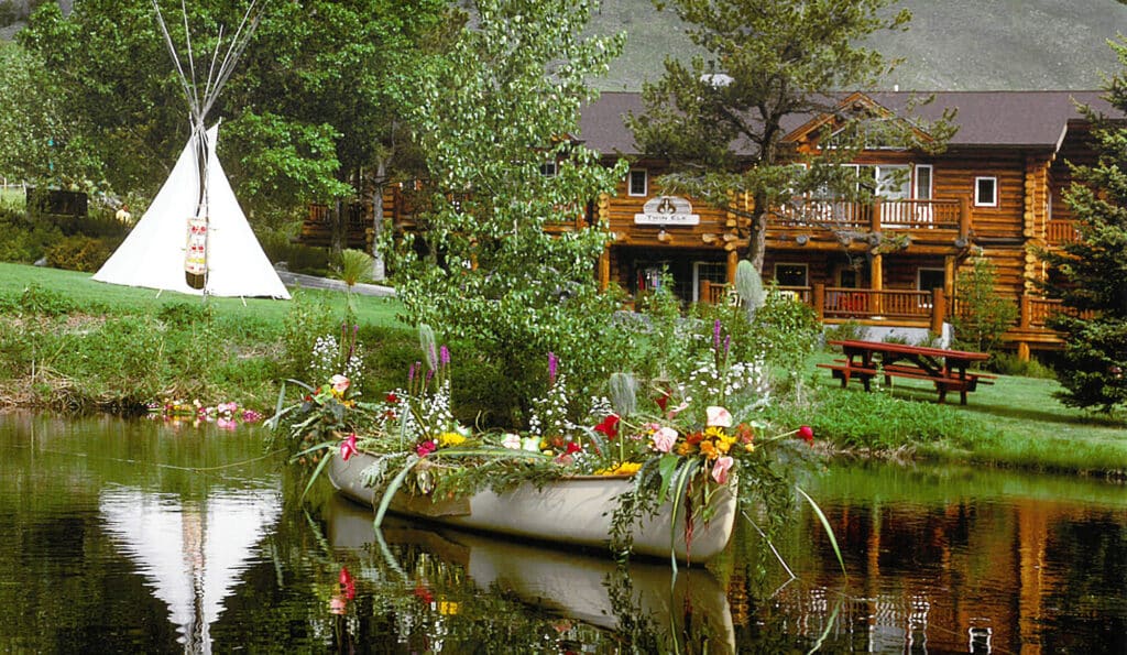 A canoe decorated with colorful flowers floats on a pond in front of a log cabin lodge, with a white teepee and green trees in the background—an inviting glimpse of recreational land for sale perfect for outdoor adventures.