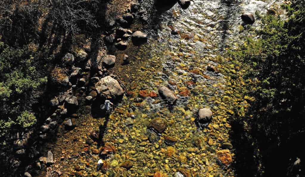 Aerial view of a rocky stream surrounded by trees and bushes, with sunlight reflecting off the water. A person stands on rocks near the left edge—an ideal spot to experience this scenic recreational land for sale.