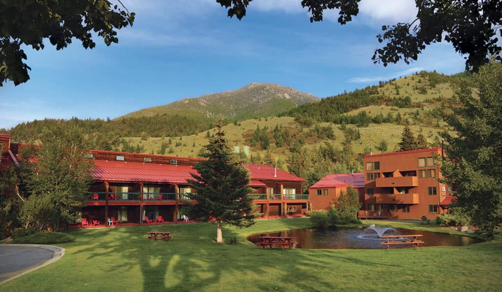 A lodge-style resort with red-roofed buildings sits on a grassy lawn with picnic tables, a small pond, and a water fountain, surrounded by trees and mountains under a blue sky—perfect as a cattle ranch or unique land for sale opportunity.