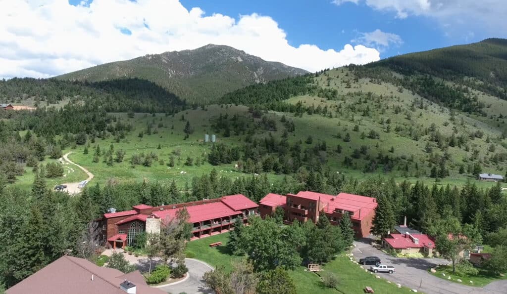 Aerial view of a large ranch for sale with red roofs, surrounded by green trees and grassy hills, set against a backdrop of tall, forested mountains under a partly cloudy sky.
