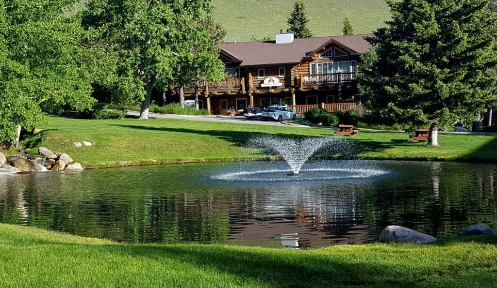 A small pond with a water fountain is surrounded by green grass and trees. In the background, a rustic wooden lodge with balconies stands near parked cars. Benches circle the pond, creating an inviting recreational land for sale.
