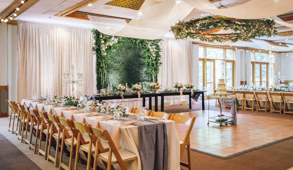 Elegant wedding reception hall with long dining tables, wooden chairs, floral centerpieces, and white linens. Ideal for celebrations near recreational land or a ranch for sale, with lush green backdrops and greenery over the dance floor.