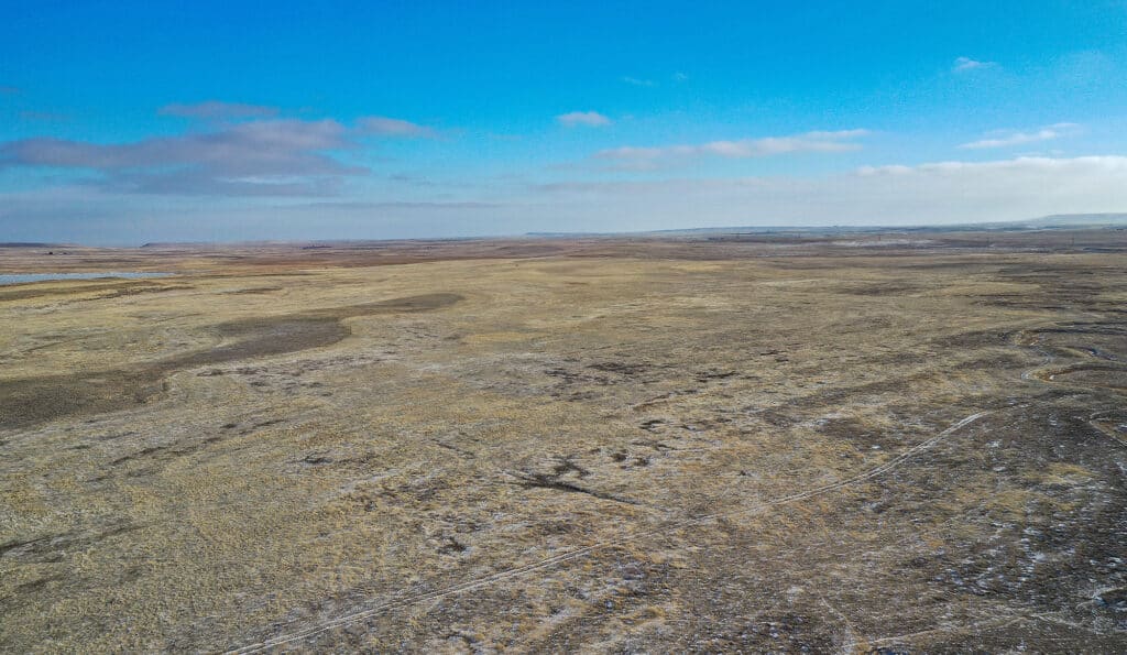 Aerial view of a vast, flat, and dry grassland landscape perfect for a cattle ranch, under a bright blue sky with scattered clouds. Distant hills are visible on the horizon.