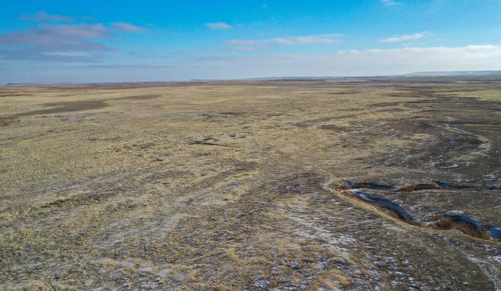 A vast, flat landscape of dry, yellow-brown grassland stretches to the horizon under a partly cloudy blue sky, with patches of snow and shallow ravines—ideal recreational land or hunting property for sale.