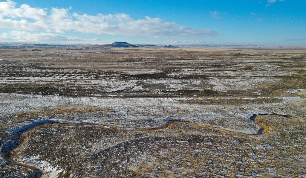 A vast, flat landscape stretches into the distance, dusted with patches of snow and dry grass—ideal recreational land or a potential cattle ranch. A small, flat-topped hill rises near the horizon under a blue sky dotted with clouds.