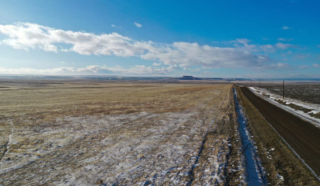 A rural, snow-dusted landscape featuring land for sale with an empty dirt road running alongside flat, open fields under a blue sky and scattered clouds. Low hills are visible on the horizon.