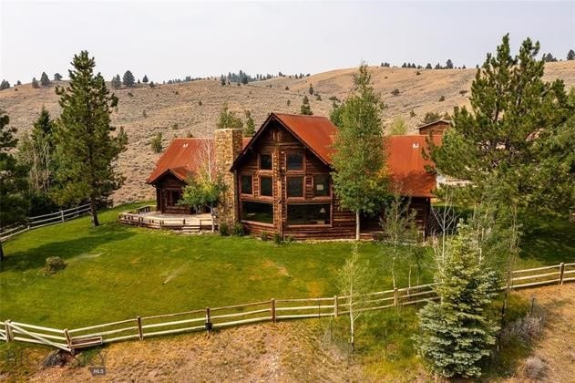 A large rustic log cabin with a red metal roof sits on a grassy plot of recreational land, surrounded by a wooden fence, trees, and rolling hills under a hazy sky.