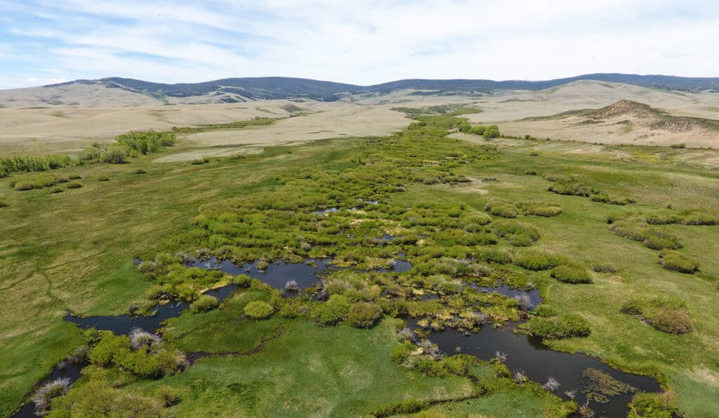 Aerial view of a lush, green wetland area with patches of water and vegetation, ideal as recreational land or hunting property, surrounded by rolling hills and distant mountains under a partly cloudy sky.