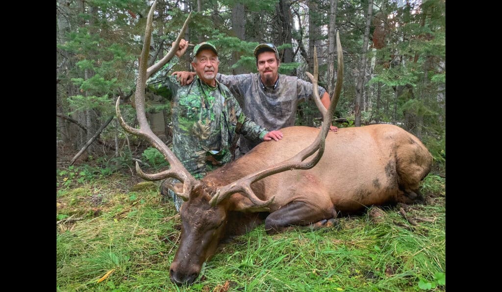 Two men in camouflage kneel in a forest, posing with a large elk on the grass. Smiling at the camera, they display their trophy amid dense green trees—highlighting the appeal of prime hunting property or recreational land.