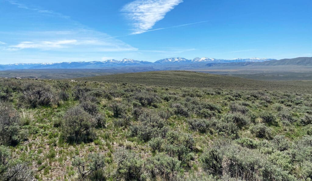 A vast sagebrush plain stretches toward distant snow-capped mountains under a clear blue sky with a few scattered clouds—perfect recreational land or hunting property, with open, sunlit views and low vegetation in the foreground.