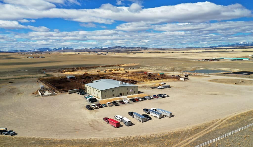 Aerial view of a rural cattle ranch surrounded by dry fields, with trucks and cars parked outside a large building, animal pens nearby, and mountains visible in the distant background under a partly cloudy sky.
