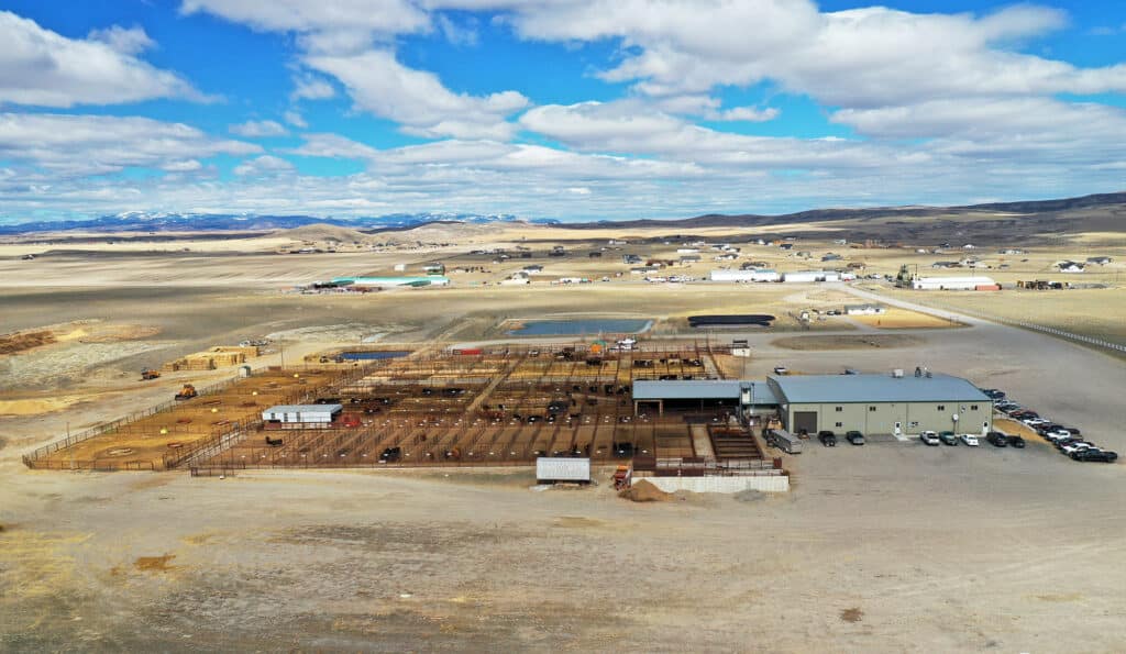 Aerial view of a large cattle ranch for sale with multiple fenced pens, adjacent buildings, parked vehicles, and open land under a partly cloudy sky. Snow-capped mountains are visible in the distance.