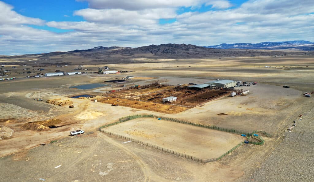 Aerial view of a cattle ranch facility in a dry, rural landscape with pens, enclosures, scattered buildings, and mountains in the background under a partly cloudy sky—ideal recreational land or land for sale.