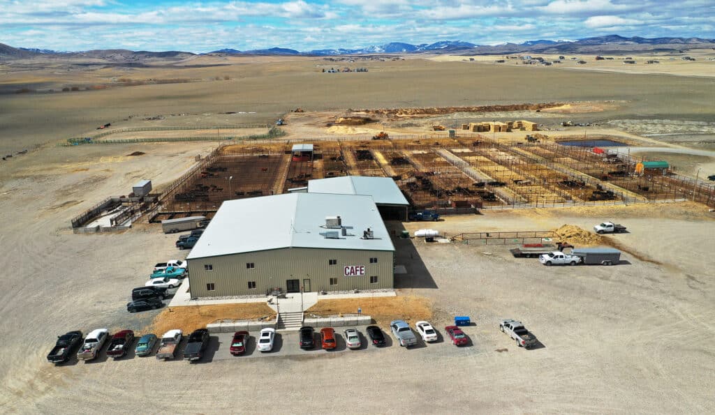 Aerial view of a rural cattle ranch with a large building labeled “CAFE,” several parked cars in front, fenced livestock pens behind, and distant mountains under a partly cloudy sky. Ideal hunting property or ranch for sale.