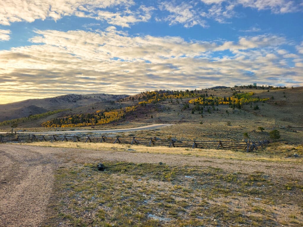 A wide, open landscape with rolling hills, scattered trees in autumn colors, a wooden split-rail fence, and a partly cloudy sky at sunrise or sunset—perfect recreational land or hunting property with grass, dirt, and sparse vegetation.