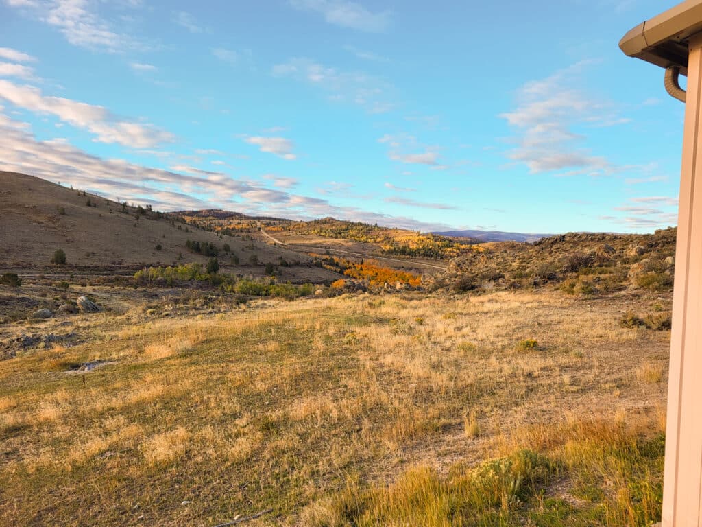 A wide, sunlit landscape with dry grass, rolling hills, scattered trees with fall foliage, and a partly cloudy sky—ideal recreational land or ranch for sale. The corner of a light-colored building is visible on the right edge.
