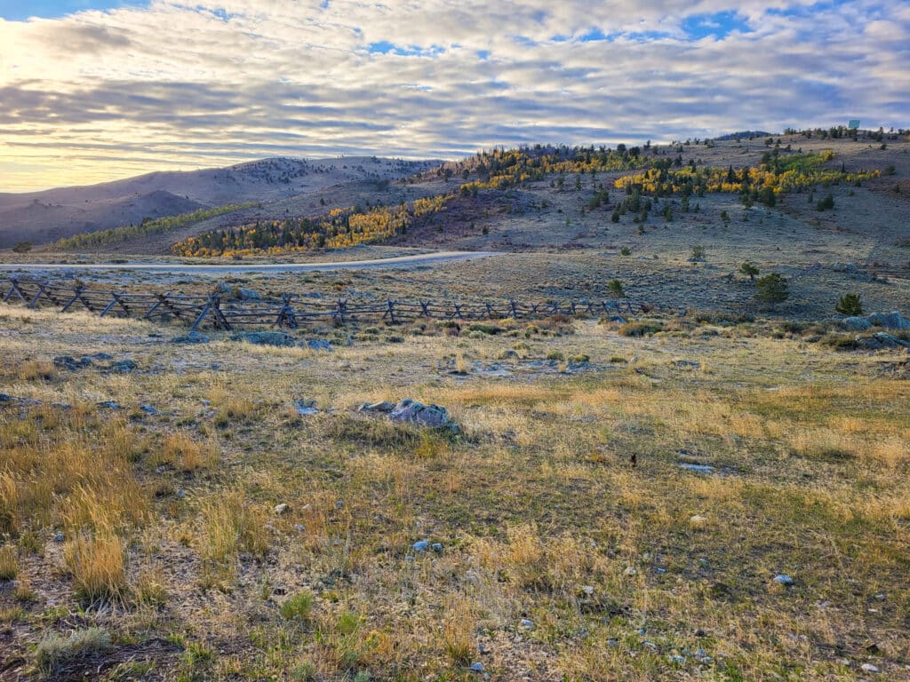 A scenic landscape of rolling hills with scattered yellow and green trees under a partly cloudy sky, a wooden split-rail fence, and dry grass in the foreground—ideal land for sale or as a hunting property, all bathed in soft sunlight.