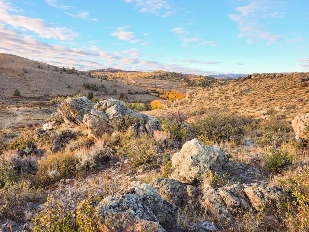 Rocky outcrops and shrubs cover a sunlit, rolling landscape with dry grass—ideal recreational land or cattle ranch territory—with sparse trees and distant hills under a blue sky. Patches of yellow foliage dot the background.