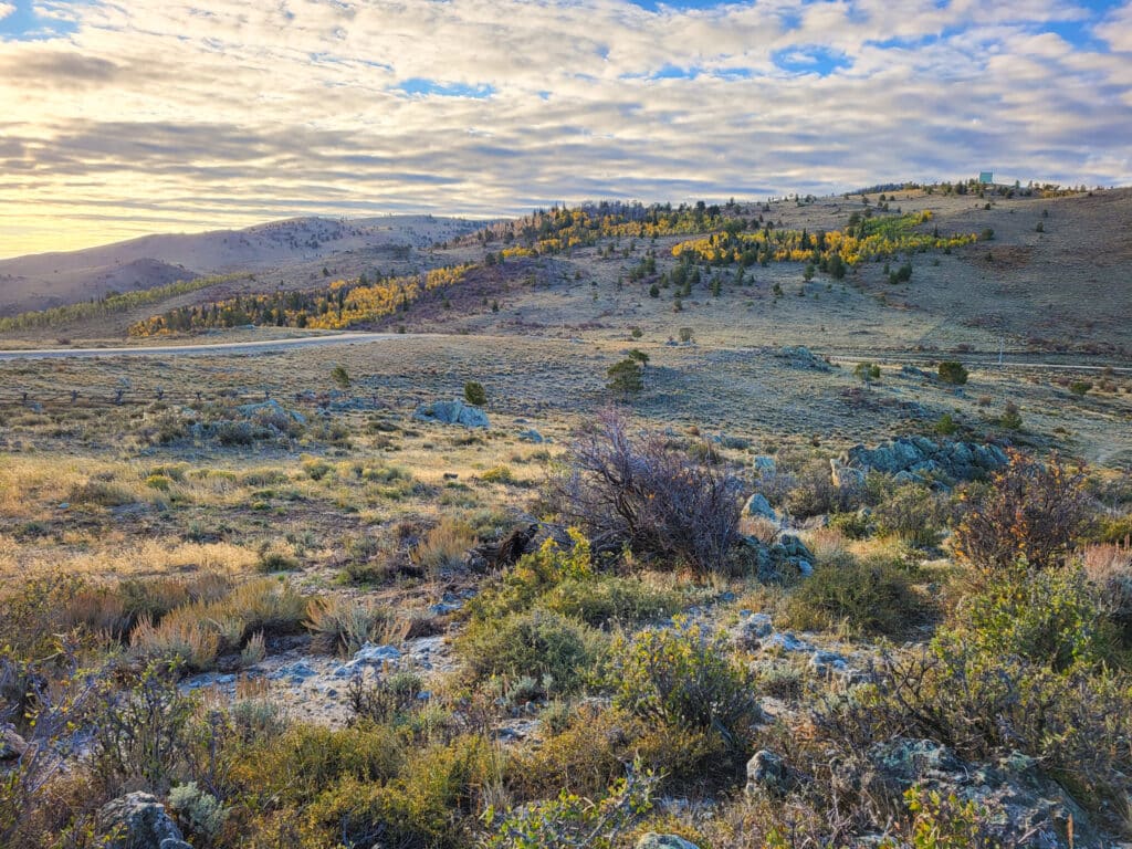 A wide, open landscape with rocky terrain, scattered shrubs, and patches of yellow autumn trees stretch across rolling hills—an ideal hunting property beneath a partly cloudy sky at sunset.